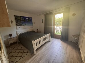 Ein Schlafzimmer mit Bett, Nachttisch, einem Gemälde, einem Fenster mit Vorhängen und Blick ins Freie.