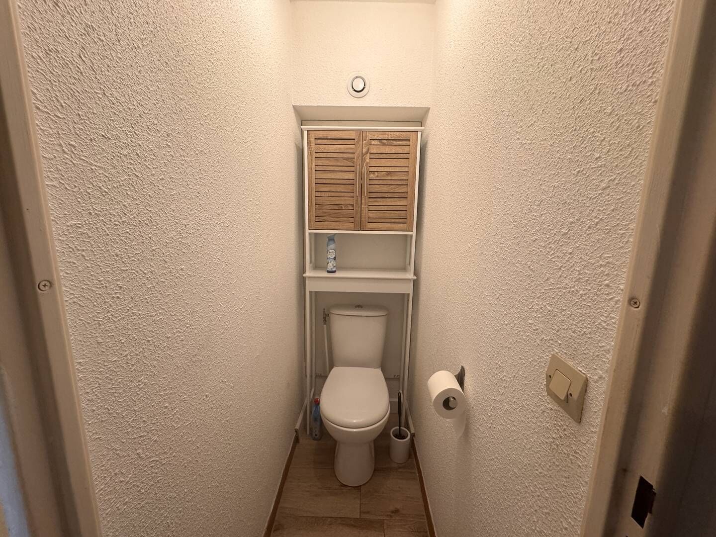 A small, narrow bathroom with a toilet, a wooden cabinet above it, and a roll of toilet paper on the wall.