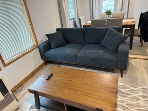 A living room with a grey sofa, a wooden coffee table, a remote control, and a dining area with a table and chairs.