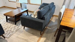Ett rum med en grå soffa, ett träbord som används som soffbord, ett matbord och en mönstrad matta.
