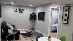 A modern living room with a sofa, a coffee table, a dining table with chairs, a TV, and a plant. - Room-cozy 2BR Suite at Winnipeg South (Winnipeg)