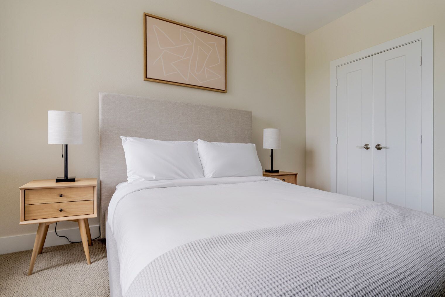 A neatly arranged bedroom with a bed, two bedside tables, a nightstand, a wardrobe, and a framed abstract artwork on the wall.