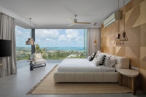 A modern bedroom with a large bed, a TV, and a view of a coastal landscape. - Villa Vie (Koh Samui)
