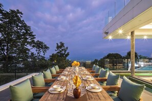 Une table à manger dressée pour un repas, avec des chaises, un vase rempli de fleurs et une vue sur le ciel et le paysage.