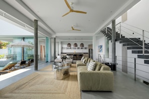 A spacious living area with a large sofa, a coffee table, and a dining area with chairs. There is a staircase with a metal railing and a modern kitchen in the background. - Villa Vie (Koh Samui)
