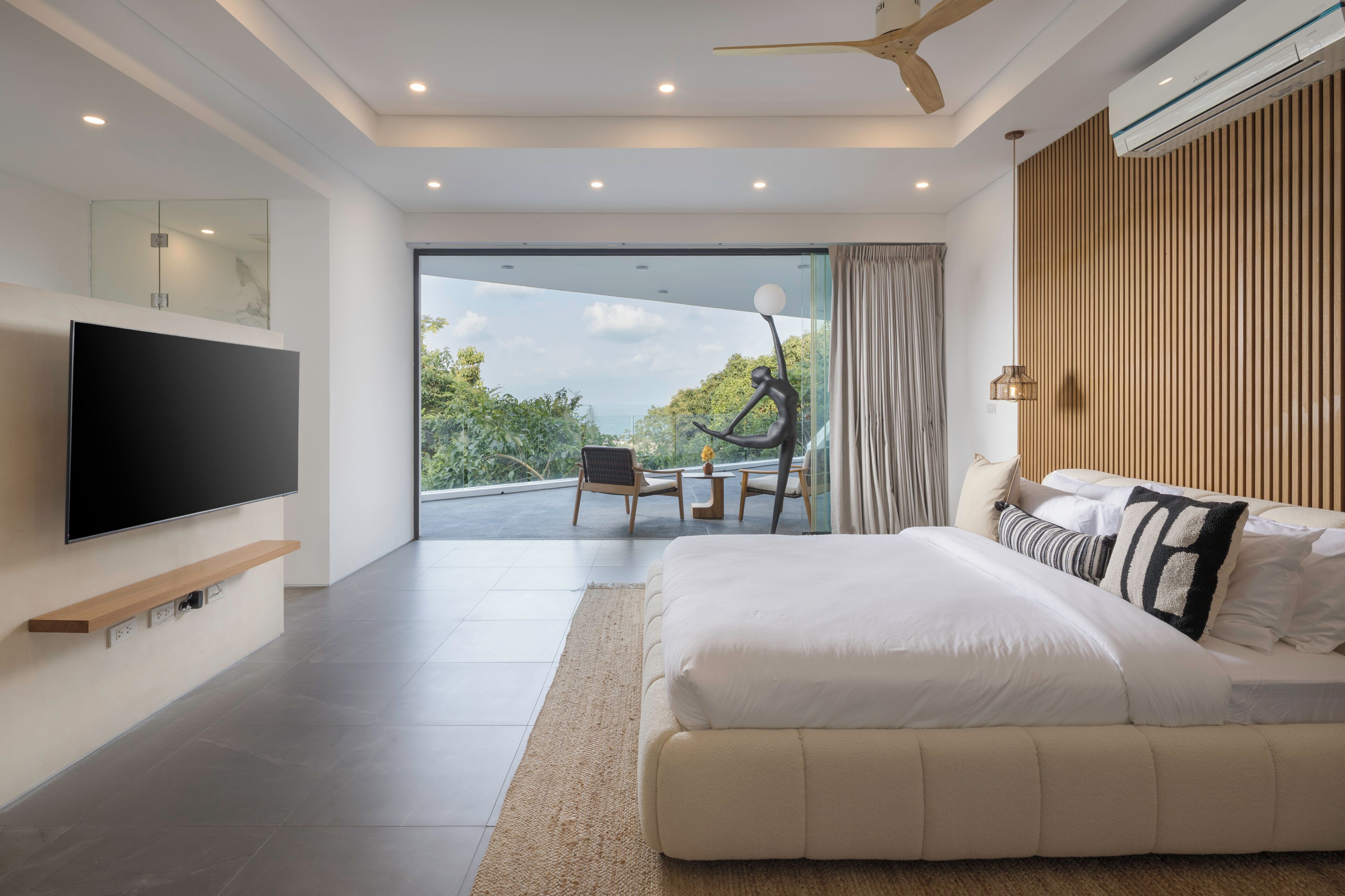 Een moderne slaapkamer met een groot bed, een flatscreen televisie, een houten plank, een plafondventilator en uitzicht op groen.