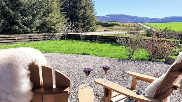 A wooden deck with two chairs and two wine glasses, overlooking a green field and a mountain range. - Nether Clashnoir Glenlivet (Ballindalloch)