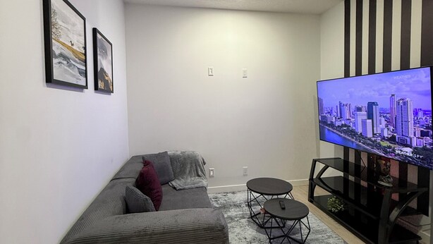 A modern living room with a grey sofa, a small round table, a flat-screen TV, and a cityscape on the screen. - Selah’s Suite - (AC+Wifi+3beds(1 King bed +2 twin sofa beds ) (Edmonton)