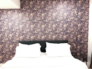 Cama com lençóis brancos, cabeceira escura e papel de parede com padrão floral.