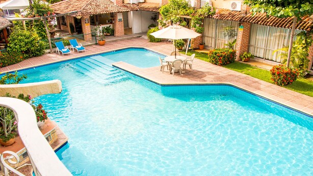 Piscina com espreguiçadeiras e uma mesa, rodeada por vegetação e um edifício de tijolo.