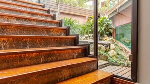 Unos escalones de madera conducen a una puerta de vidrio, con vista a un jardín y un edificio al fondo.