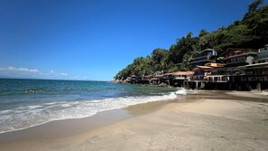 A beach with a clear blue sky, calm sea, and houses built on stilts along the shoreline. - Manoir Oasis: 5 spacious suites and exclusive beach access (Guarujá)