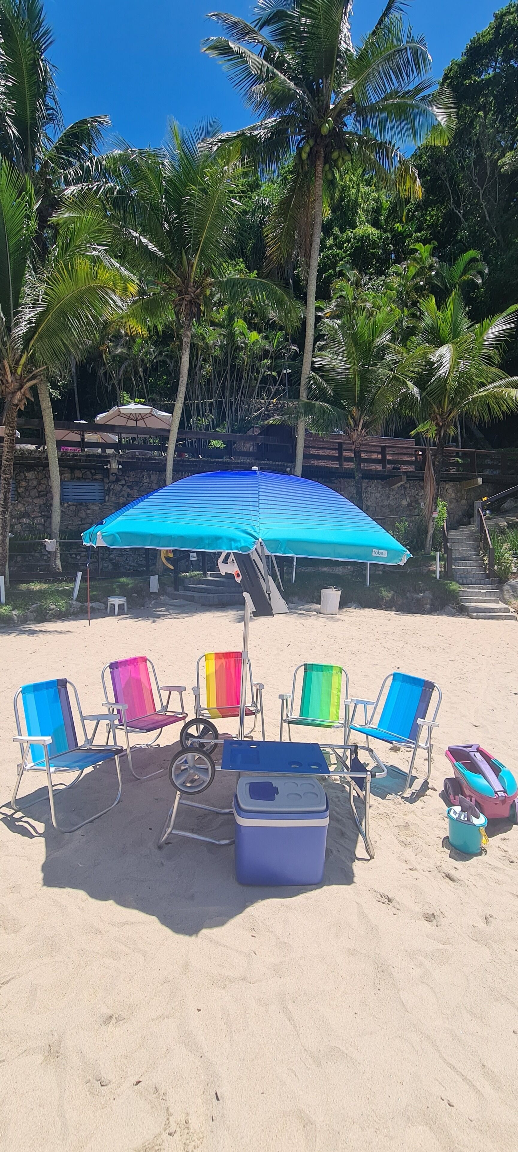 Une plage avec un parasol bleu, des chaises de plage colorées et une glacière sur le sable.