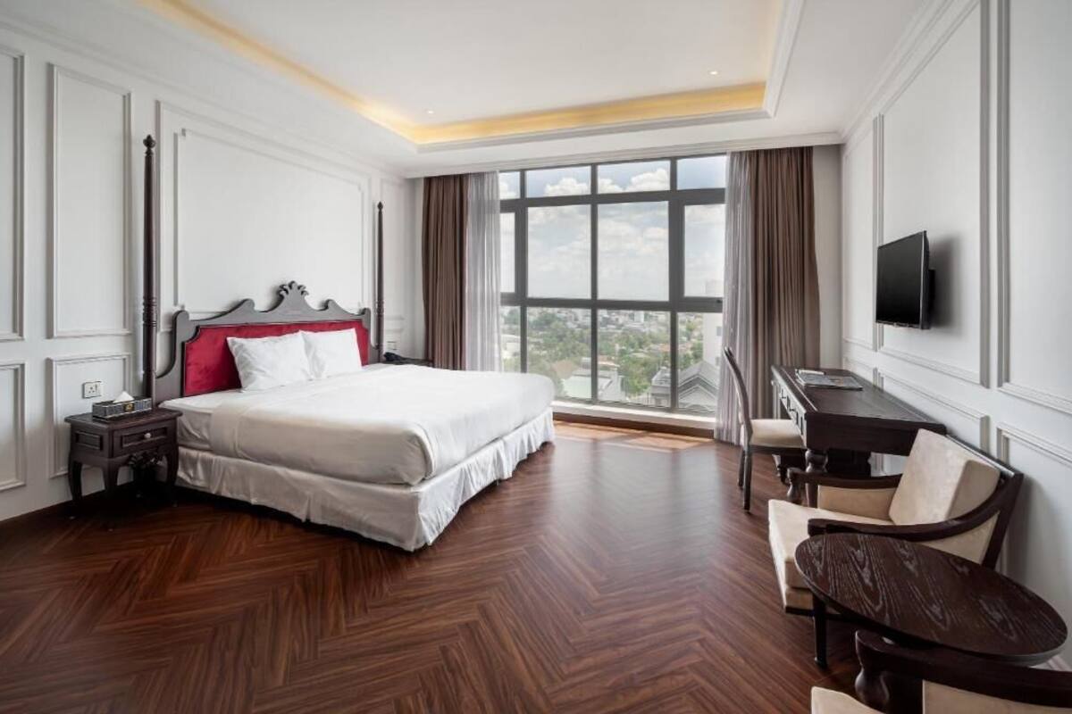 A hotel room with a large bed, a desk with a chair, a television, and a large window with a city view.