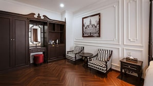 Um quarto com um guarda-roupa grande de madeira escura, um banquinho vermelho, duas poltronas listradas, uma mesa redonda pequena e um quadro na parede.