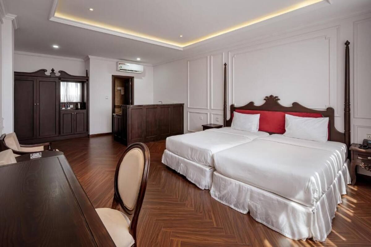 A hotel room with a double bed, a wooden desk, a chair, a wardrobe, and an air conditioning unit.