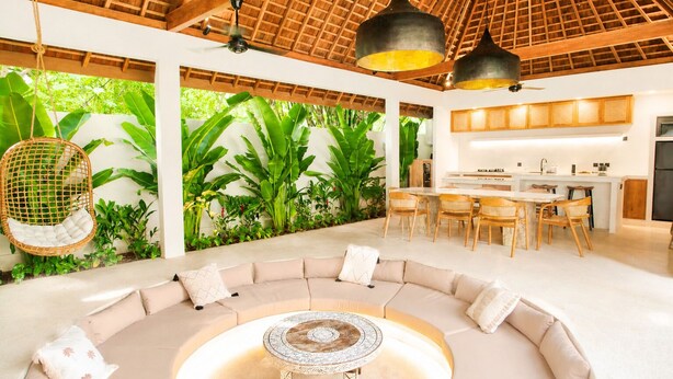 Una cocina moderna con una isla central, un comedor con sillas de mimbre, una zona de estar circular con cojines y un ventilador de techo.
