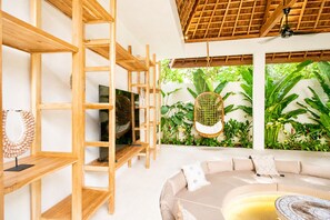A modern living room with a TV, wooden shelves, a sofa, and a large window with a view of greenery. - Villa Toro - Bingin Bali (Pecatu)