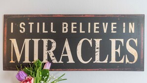 Et skilt med teksten 'I STILL BELIEVE IN MIRACLES' monteret på en væg.