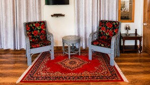 Una habitación con dos sillones de mimbre con tapicería floral, una mesita redonda pequeña, un tapete estampado rojo, un televisor de pantalla plana montado en la pared y una mesita de noche de madera con una lámpara.