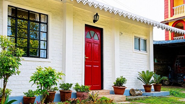 Un edificio blanco con puerta roja, plantas en macetas y un ventanal pequeño.