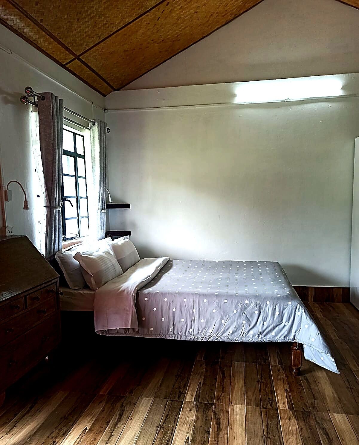A single bed with a grey bedspread, a wooden dresser, a window with curtains, and a wooden ceiling.