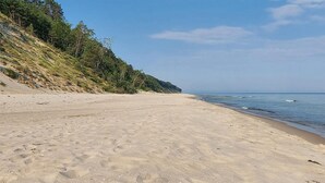 A sandy beach with gentle waves, a clear sky, and a forested hillside. - Lovely home in Ko1czewo (Ko1czewo)