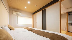 A hotel room with two beds, a wardrobe, a window with blinds, and an air conditioning unit.