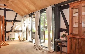 Una stanza con soffitto in legno, ampie finestre con tende, un tavolino con un vaso e una porta in legno.