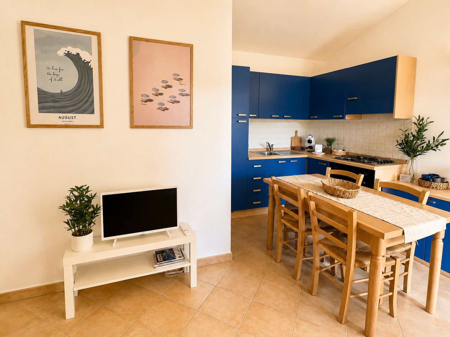 A modern kitchen with blue cabinets, a dining table with chairs, a TV on a stand, and two framed pictures on the wall.