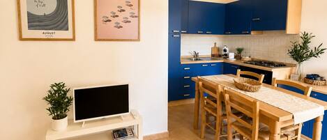 A modern kitchen with blue cabinets, a dining table with chairs, a TV on a stand, and two framed pictures on the wall.