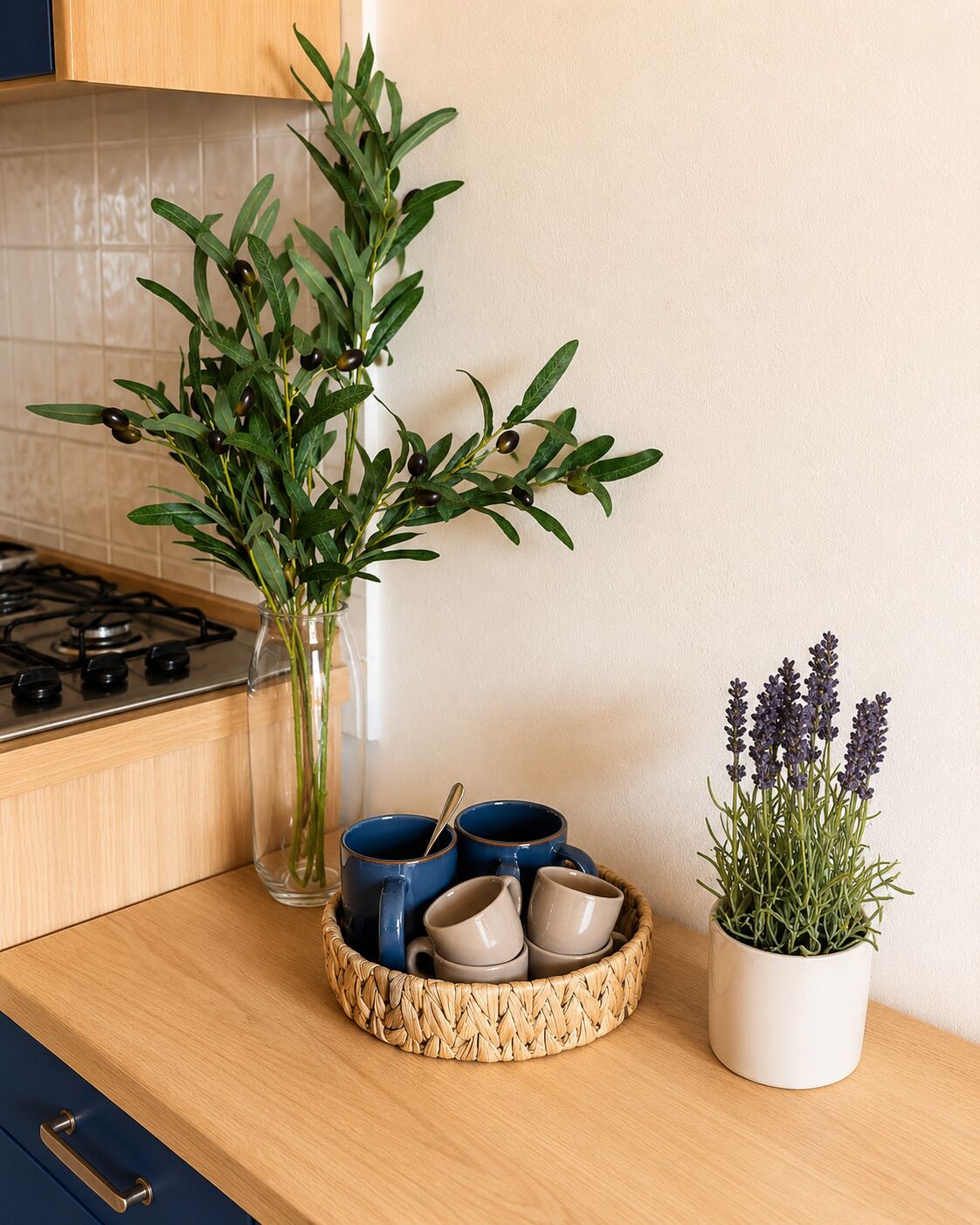 A kitchen counter with a clear glass vase containing green leaves, a woven tray with blue and beige mugs, and a potted lavender plant.