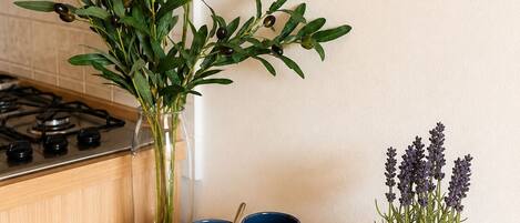 A kitchen counter with a clear glass vase containing green leaves, a woven tray with blue and beige mugs, and a potted lavender plant.