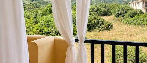 Ein Balkon mit weißen Vorhängen, einem Blumentopf und Blick auf eine grüne Landschaft mit Hügeln.