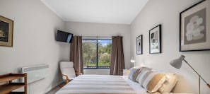 Un dormitorio con cama, una silla, un escritorio, un televisor y una ventana con cortinas.