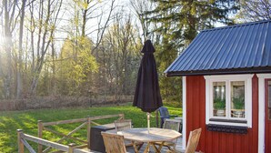 Eine Holzterrasse mit Tisch und Stühlen, ein rotes Gebäude mit blauem Dach und ein bewaldeter Hintergrund.