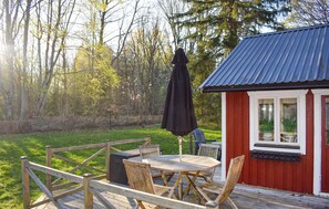 Eine Holzterrasse mit Tisch und Stühlen, ein rotes Gebäude mit blauem Dach und ein bewaldeter Hintergrund.