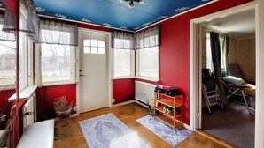 A room with a red wall, white door, and windows with white blinds. There is a wooden floor, a bench, and a shelf with shoes.