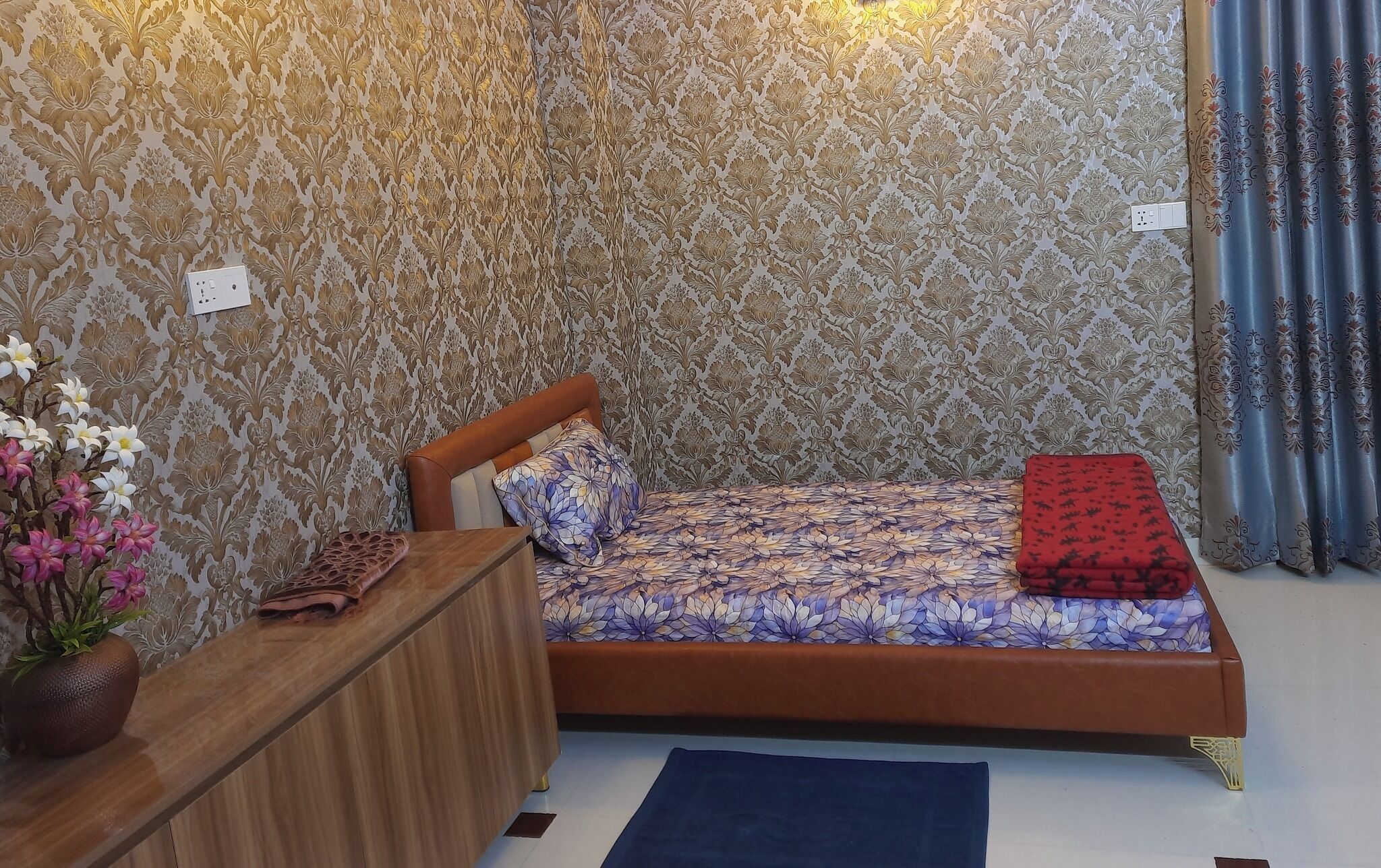 Un dormitorio con una cama, un aparador de madera, un florero con flores y papel tapiz estampado.