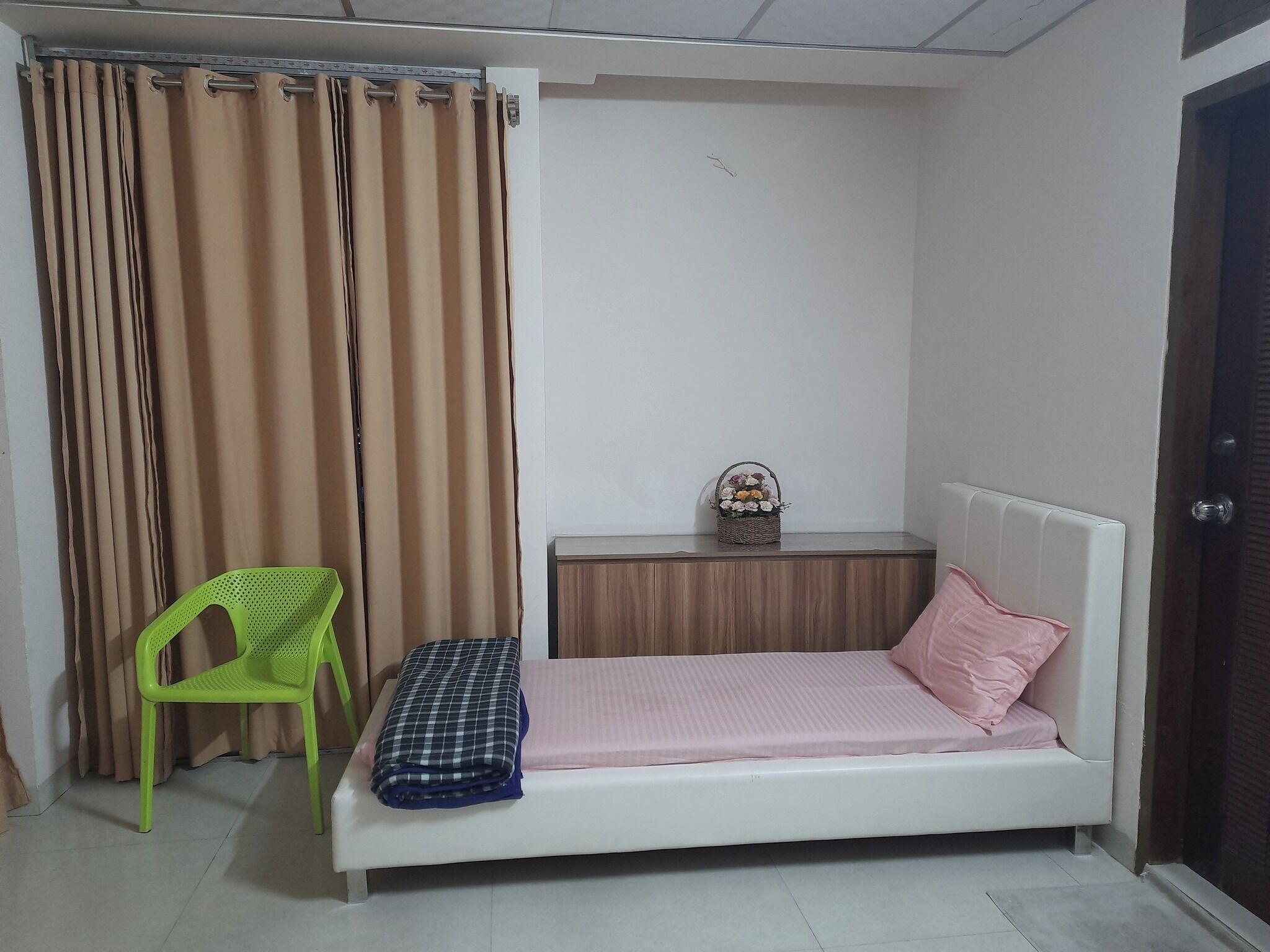 A small, neatly arranged bedroom with a single bed, a green chair, a bedside table with a basket, and a curtain.