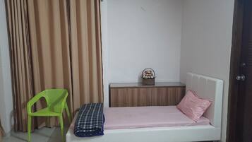 A small, neatly arranged bedroom with a single bed, a green chair, a bedside table with a basket, and a curtain.