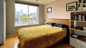 Ein Schlafzimmer mit Bett, Deckenventilator, Fenster mit Stadtblick, Bücherregal und Backsteinwand.