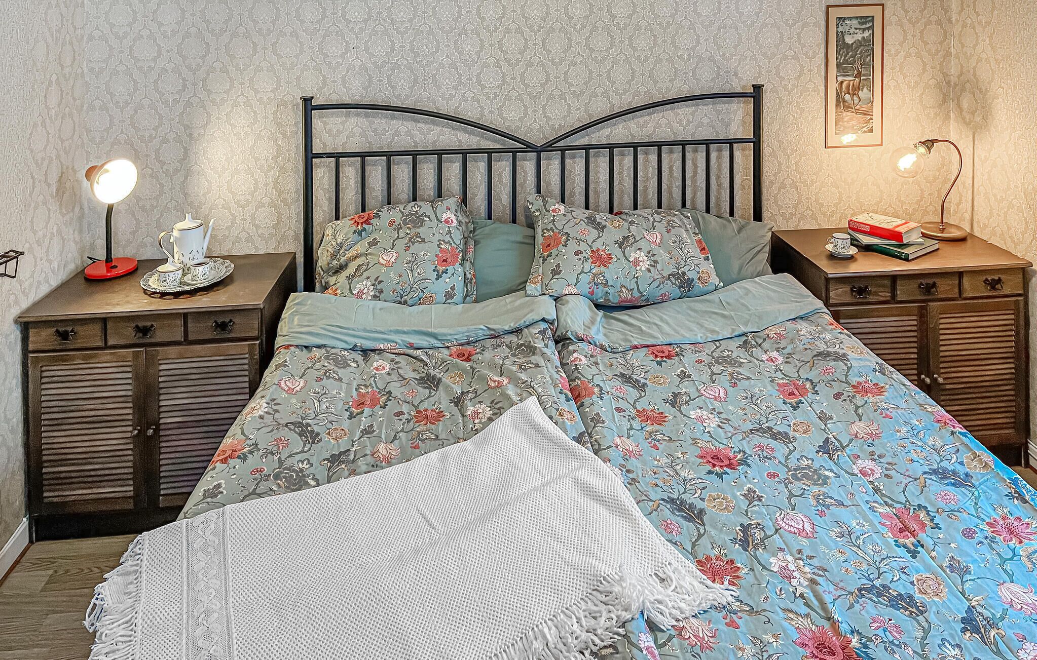 Un lit avec une literie à motifs floraux, deux tables de chevet avec des lampes, un tableau encadré au mur et une petite étagère avec des livres et une tasse.