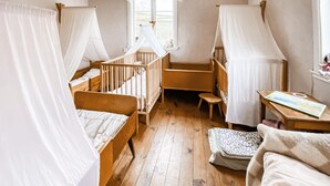 Ein Zimmer mit zwei hölzernen Babybetten, einem kleinen Holztisch, einem Fenster mit Blick und einem weißen Vorhang.