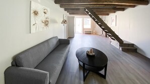 A spacious room with a grey sofa, a round black table with candles, wooden flooring, a staircase, and a window allowing natural light.