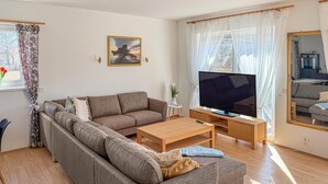 Ein Wohnzimmer mit einem Ecksofa, einem Fernseher auf einem Holzschrank, einem kleinen Beistelltisch und einem Fenster mit Vorhängen.