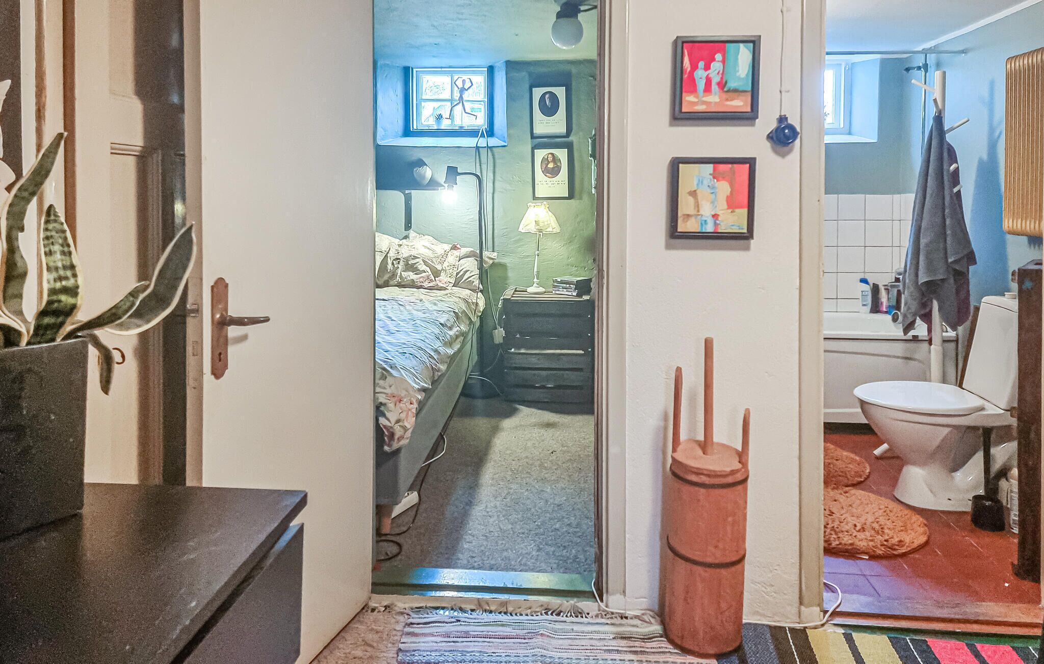 A narrow doorway leading into a bedroom with a bed, bedside table, lamp, and wall art.