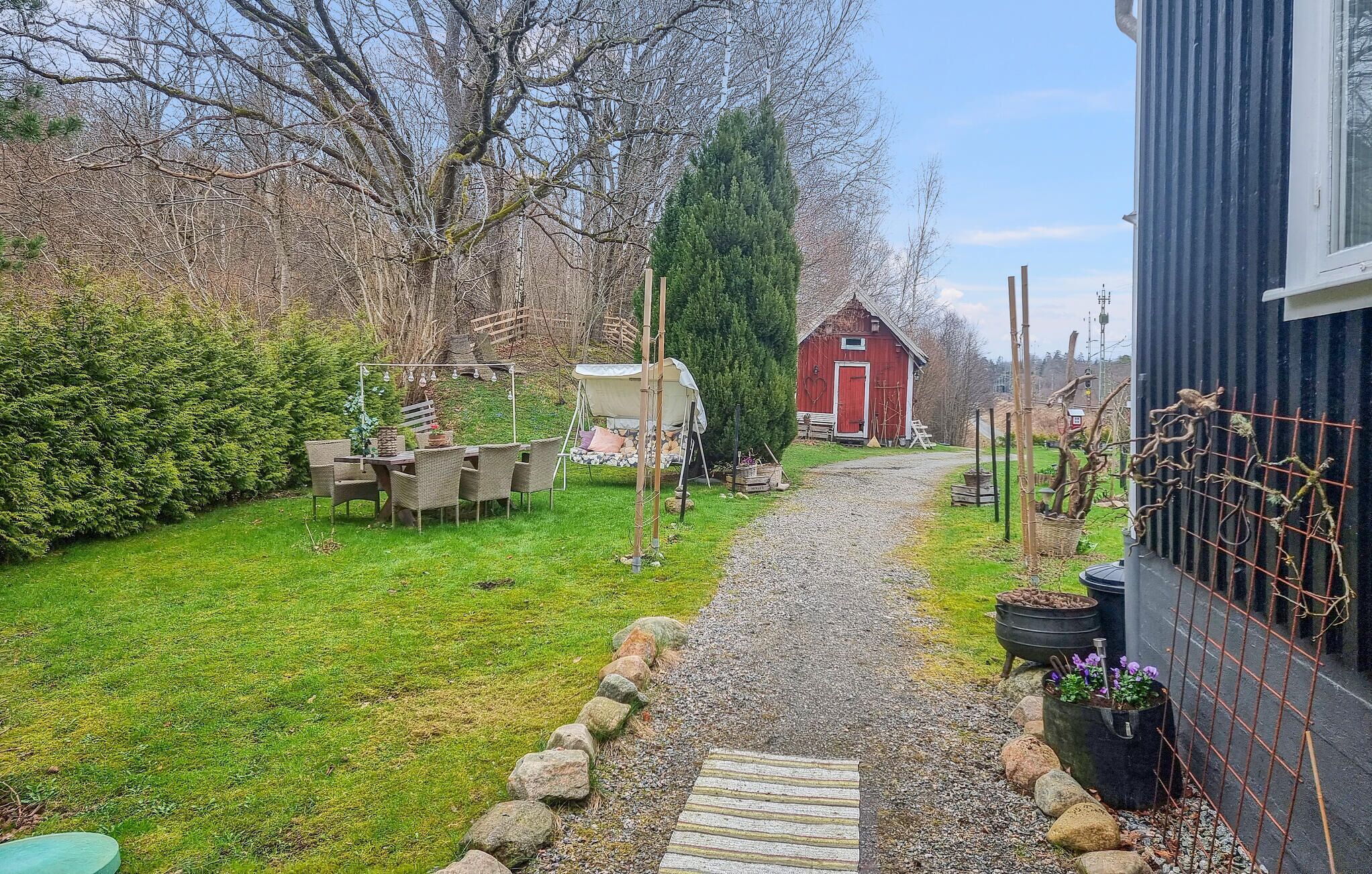 A gravel path leading to a red barn, surrounded by greenery and a fenced area with plants.