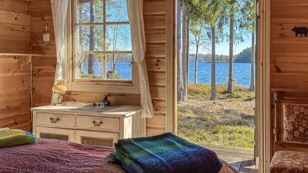 A wooden cabin room with a bed, a dresser, a window with curtains, and a view of a lake. - Awesome home in Grythyttan with lake view (Grythyttan)