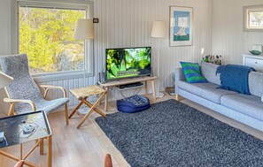 Un salon comprenant un téléviseur, un canapé, un fauteuil, une petite table et une fenêtre donnant sur des arbres.